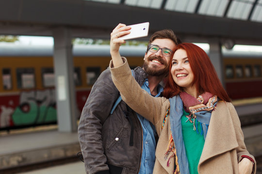 Couple of hipster travelers photographing a selfie with a smartphone in a train station. Travel concept. Mobile photography. Autumn time. Redhair woman and man in glasses