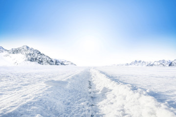 Fototapeta na wymiar Winter Background of foreground of road covered with snow and ice. In the background, the arctic landscape of the mountains with the beautiful blue sky.