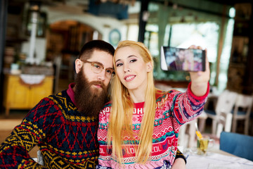 Close up of young pretty hipster love couple in a sweater hugging and taking a selfie at home for Christmas holidays.