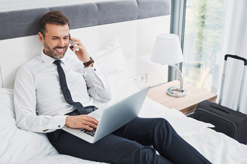 Handsome businessman making phonecall while sitting on hotel room bed and using laptop during...