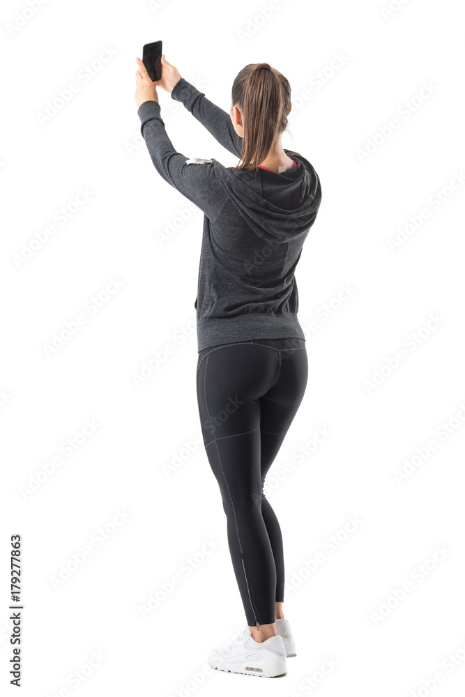 Wall mural Rear view of sporty athletic woman taking selfie while holding cellphone in both hands. Full body length portrait isolated on white background.