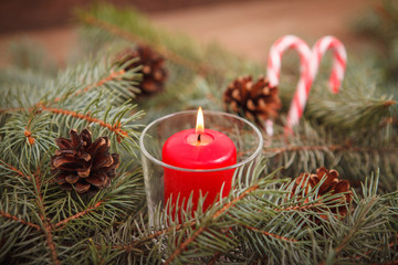 Obraz na płótnie Canvas New Year composition, burning red candle in a candlestick on spruce branches with cones on a wooden background with candy-cane. Close-up. Christmas.