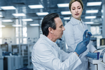 Fototapeta premium Professional lab. Smart experienced male scientist sitting at the table and looking at the test tube while working on his research