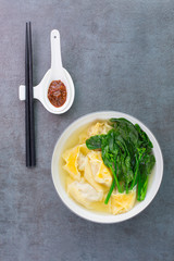 A bowl of Cantonese wonton with soup and Gai lan. A spoon with chili sauce and chopsticks next to the bowl 