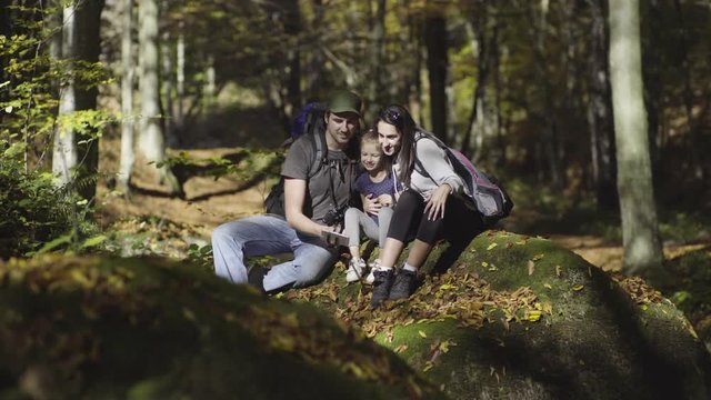 Happy family, man, woman and little child sitting on stone with yellow leaves in wood, hiker photographing with android phone on stick, fall, sunny day, shallow depth of field, concept hiking, travel