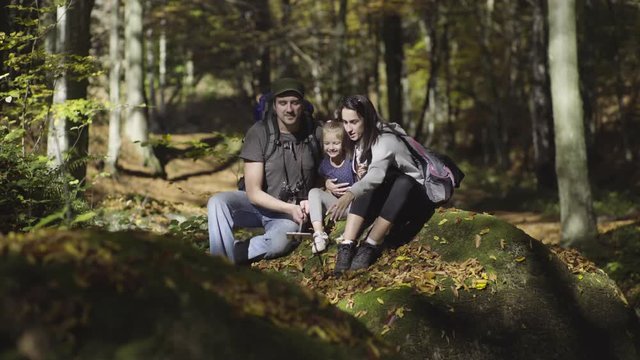 Happy Caucasian young couple and little girl sitting on rock covered with yellow leaves in forest and take photo with cell phone on stick, fall, sunny day, shallow depth of field, concept hiking,