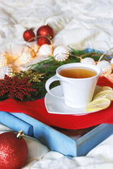 Breakfast with a cup tea, a biscuit on a tray on the bed on a white blanket in the winter morning decorated with Christmas decorations, spruce branches with luminous garlands, concept holiday