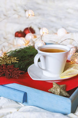Breakfast with a cup tea, a biscuit on a tray on the bed on a white blanket in the winter morning decorated with Christmas decorations, spruce branches with luminous garlands, concept holiday
