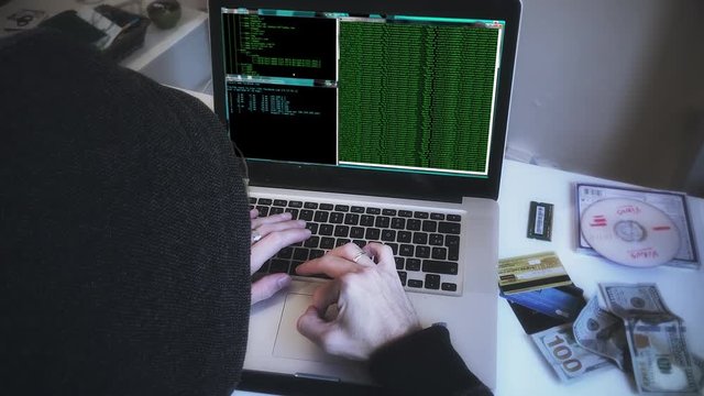 Zoom In Anonymous Hacker, Cyber Attack Crime Scene. Hooded person typing in a laptop next to stolen itens, simulating a hacker crime scene