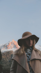 Beautiful Cacasian female wearing brown coat and hat posing against Mont Blanc massif during sunset