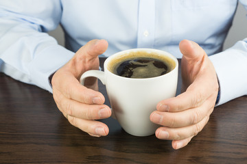 Man sitting at a brown wooden table holding a cup of hot coffee in his hands