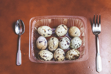 Quail eggs lay in transparent box. Near fork, spoon