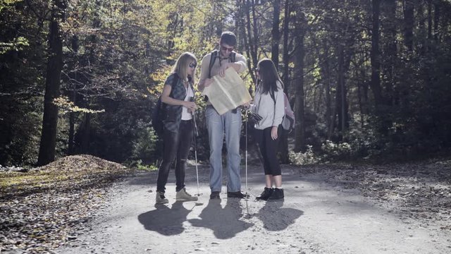 Group of hikers, Caucasian young man, woman and girl standing on the mountain path, looking a geographical map than continue to hiking on dirt road through the forest trees, backlight, autumn season