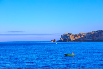 Traditional fishing boat sails in the calm waters in front of main beach of Nazare in Central Portugal. The high cliffs of Nazare with fortress and its lighthouse on background. Copy space.