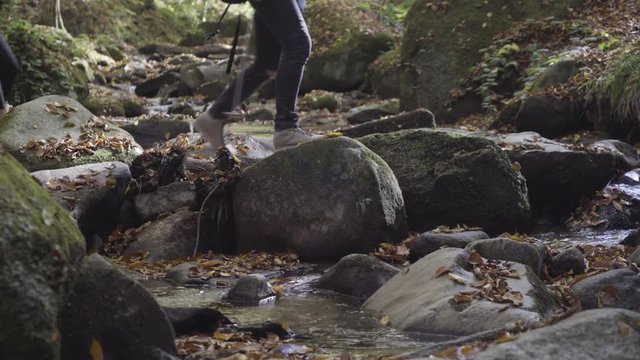 Group of hikers crossing the mountain stream over the rocks at autumn season, legs close up, creek rapids splashes, yellow fallen leaves covered environment, concept hiking, travel, symbol