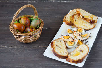 Easter wicker basket with colored eggs and sliced Easter bread in white plate on grey wooden board.