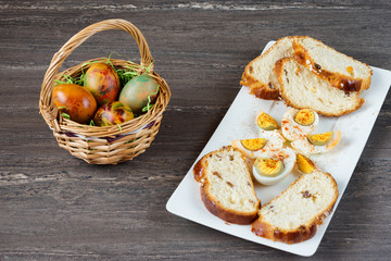 Easter wicker basket with colored eggs and sliced Easter bread in white plate on grey wooden board.