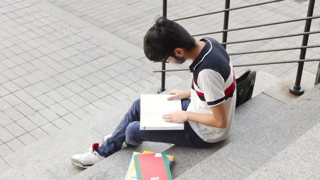 Portrait of a happy male asian student sitting on stairs and reading book