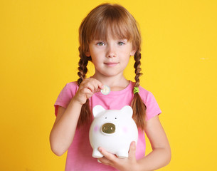Fototapeta na wymiar Cute little girl putting coin in piggy bank on color background