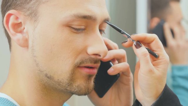Busy showman talks on phone during visagist doing him a makeup