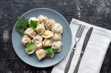 Boiled dumplings (pelmeni) with fresh parsley and dill on dark slate backdrop. Silverware on a white textile. Top view 