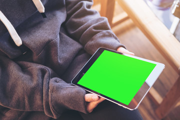 Mockup top view image of a woman sitting cross legged and holding black tablet pc with blank green screen on thigh and wooden floor background