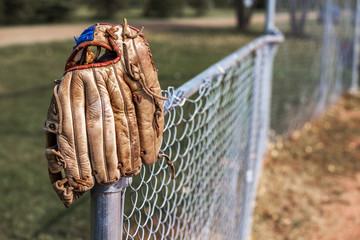 An old scuffed leather baseball glove hanging on the post of a chain link fence