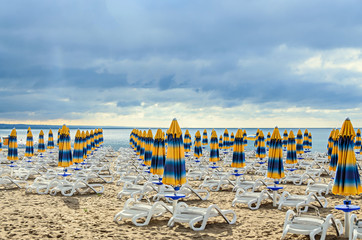 Colored stripped sun umbrellas, golden beach sand and sunbeds, sea water and clouds sky