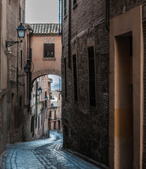 A narrow street with an arch connecting two opposite buildings. The street runs downwards opening a view to thw mountains. Invitation to travel