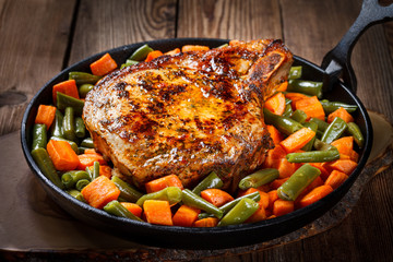 Pork grilled steak with carrots and green beans in a cast-iron frying pan on a natural wooden background.