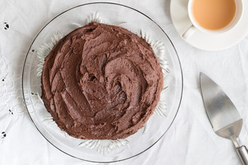 Chocolate cake whole on glass serving dish, metal server and cup of tea on white table cloth - Top view image