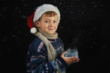 Boy in a sweater and Santa hat holding in hands a hot Cup on a dark background .