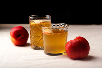 Two vintage glasses with apple cider on black background. Christmas beverages concept. Two red apples and rosemary sprig aside.  Warm backlight. Horizontal composition. Side view.