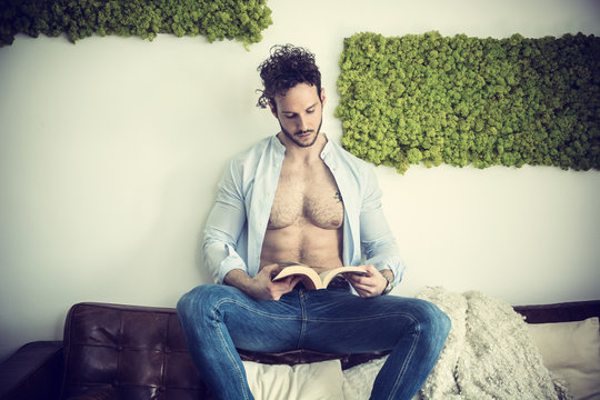 Shirtless sexy male model lying alone on couch in his living-room, looking at camera with a seductive attitude, reading a book,, with shirt open on muscular chest and torso