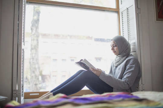 Muslim woman wearing a hijab reading on a window sill