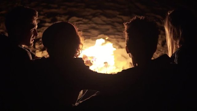 Back view of diverse group of people sitting together by the fire late at night and embracing each other. Cheerful friends talking and having fun together