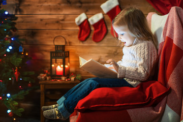 girl at home with a christmas tree, gifts reading a book sitting in a red armchair