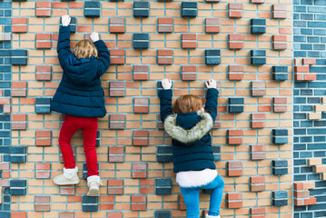 Young girls climbing a brick wall in autumn. Two kids with red hair wearing winter jackets outdoors on playground. Perfect for family blogs, outdoor business themes, urban concepts