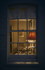 Looking through a window at night from outside into a rustic cottage with lamp