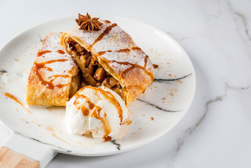 Home autumn, summer baking, puff pastries. Apple strudel with nuts, raisins, cinnamon and powdered sugar. On white marble table. Sliced, with ice cream and caramel topping. Copy space