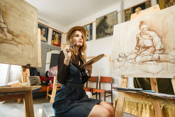 Female artist wears hat posing near picture indoor the studio