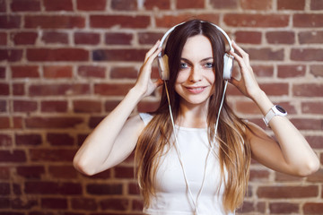 Caucasian hipster girl, with long straight hair and white headphones around her neck, smiles crossing her arms before the brick wall, isolated