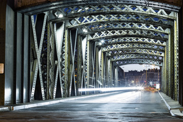 Asphalt road under the steel construction of a bridge in the city. Night urban scene with car light trails in the tunnel. Toned