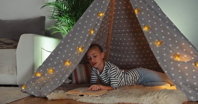 Girl relaxing in wigwam. Child reading book and smiling at camera