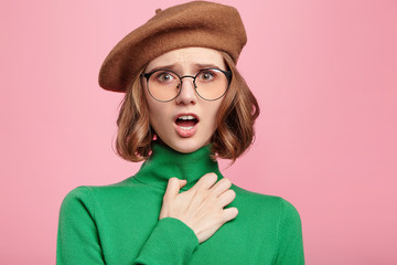 Adorable female model with bated breath, looks scared at camera, keeps hand on chest, afraid of big dog on street, wears beret and green turtleneck sweater. Emotional beautiful woman indoors