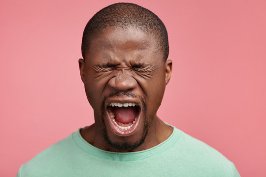 Emotional African American male closes eyes in despair, opens mouth, yells loudly, being scared of horror film. Frightened dark skinned man afraid of something, isolated over pink background