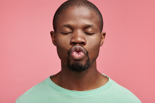 Horizontal portrait of dark skinned man roundes lips as going to kiss girlfriend, closes eyes with pleasure, poses in pink studio. Bearded African American young male makes grimace, stands alone