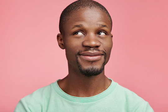 Isolated shot of pleasant looking male with dark skin has moony expression, looks aside up, dreams about something pleasant, isolated over pink background. Happiness and positiveness concept