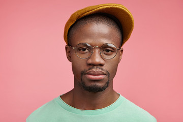 Strict serious man with dark healthy skin, wears trendy cap and glasses, has confident expression, specific appearance, looks severe as tries to look like boss. People, facial expressions concept