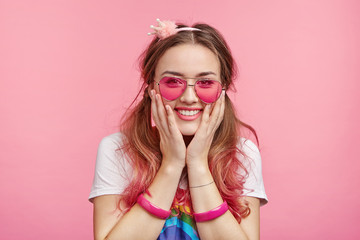 Joyful coquette female expresses positive emotions, keeps hands on cheeks, smiles happily, can`t believe in success, isolated over pink background. Delightful girl with make up, in trendy sunglasses